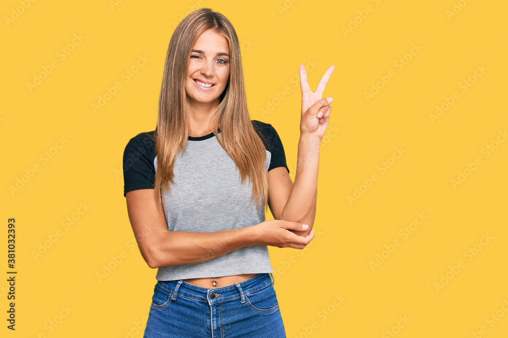 Young blonde woman wearing casual clothes smiling with happy face winking at the camera doing victory sign. number two. Young blonde woman wearing casual clothes smiling with happy face winking at the camera doing victory sign. number two.