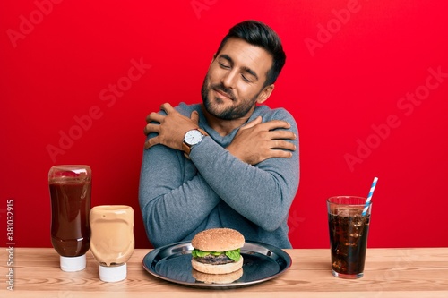 Handsome hispanic man eating a tasty classic burger and soda hugging oneself happy and positive, smiling confident. self love and self care