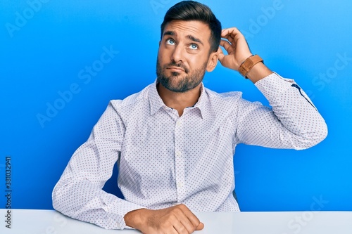 Handsome hispanic man wearing business clothes sitting on the table confuse and wondering about question. uncertain with doubt, thinking with hand on head. pensive concept. photo