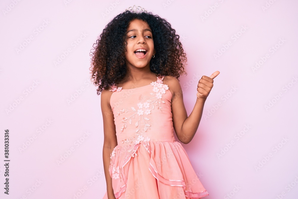 African american child with curly hair wearing princess crown pointing thumb up to the side smiling happy with open mouth African american child with curly hair wearing princess crown pointing thumb up to the side smiling happy with open mouth