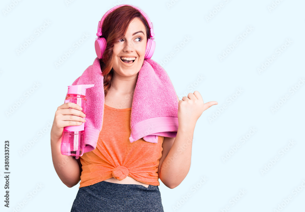 Young beautiful woman wearing sportswear and headphones holding bottle of water pointing thumb up to the side smiling happy with open mouth Young beautiful woman wearing sportswear and headphones holding bottle of water pointing thumb up to the side smiling happy with open mouth