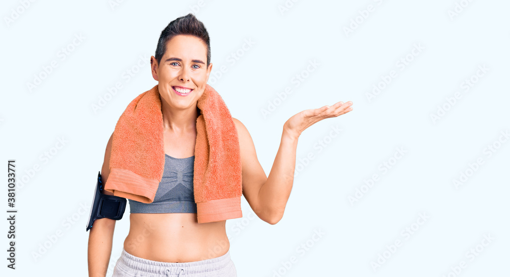 Young woman with short hair wearing sportswear and towel using smartphone smiling cheerful presenting and pointing with palm of hand looking at the camera. Young woman with short hair wearing sportswear and towel using smartphone smiling cheerful presenting and pointing with palm of hand looking at the camera.