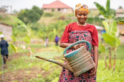 beautiful African lady with head scarf, plastic container-landscape image of Black woman in a greenfield with pretty smile-farming concept