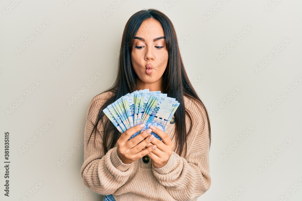 Young beautiful hispanic girl holding south african 100 rand banknotes making fish face with mouth and squinting eyes, crazy and comical. Young beautiful hispanic girl holding south african 100 rand banknotes making fish face with mouth and squinting eyes, crazy and comical.