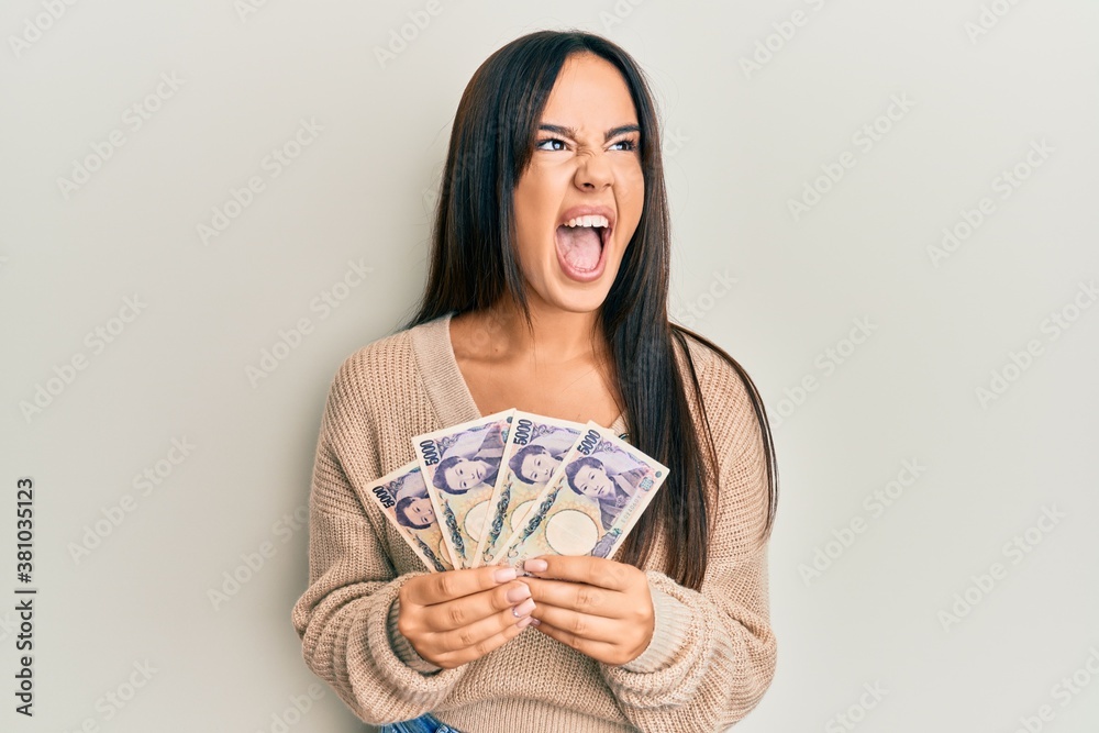 Young beautiful hispanic girl holding japanese yen banknotes angry and mad screaming frustrated and furious, shouting with anger. rage and aggressive concept. Young beautiful hispanic girl holding japanese yen banknotes angry and mad screaming frustrated and furious, shouting with anger. rage and aggressive concept.