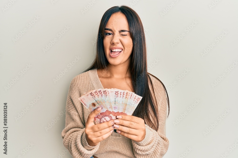 Young beautiful hispanic girl holding 10 colombian pesos banknotes winking looking at the camera with sexy expression, cheerful and happy face. Young beautiful hispanic girl holding 10 colombian pesos banknotes winking looking at the camera with sexy expression, cheerful and happy face.
