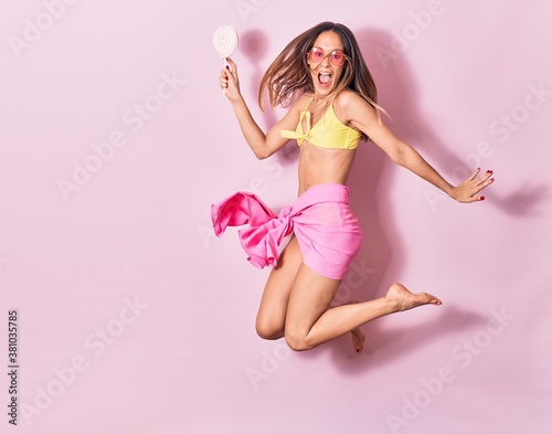 Young beautiful girl wearing bikini and sunglasses smiling happy. Jumping with smile on face holding lollipop over isolated pink background