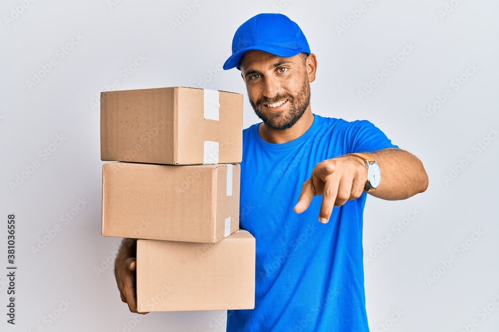 Handsome man with beard wearing courier uniform holding delivery packages pointing to you and the camera with fingers, smiling positive and cheerful Handsome man with beard wearing courier uniform holding delivery packages pointing to you and the camera with fingers, smiling positive and cheerful