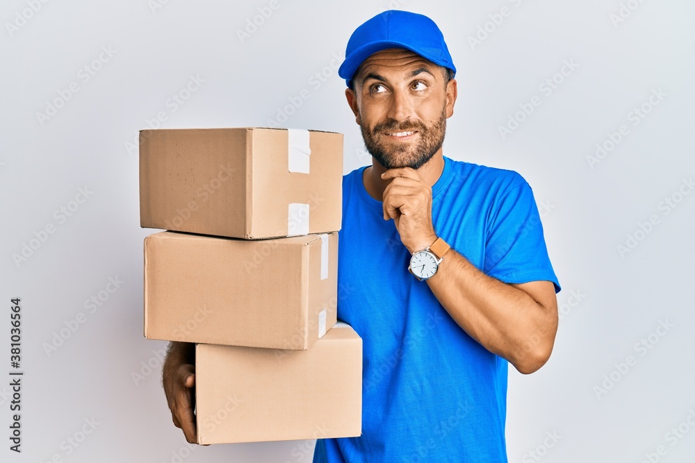 Handsome man with beard wearing courier uniform holding delivery packages with hand on chin thinking about question, pensive expression. smiling and thoughtful face. doubt concept. Handsome man with beard wearing courier uniform holding delivery packages with hand on chin thinking about question, pensive expression. smiling and thoughtful face. doubt concept.