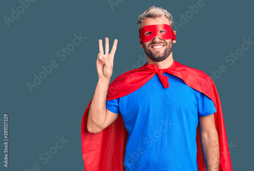 Young blond man wearing super hero custome showing and pointing up with fingers number three while smiling confident and happy.