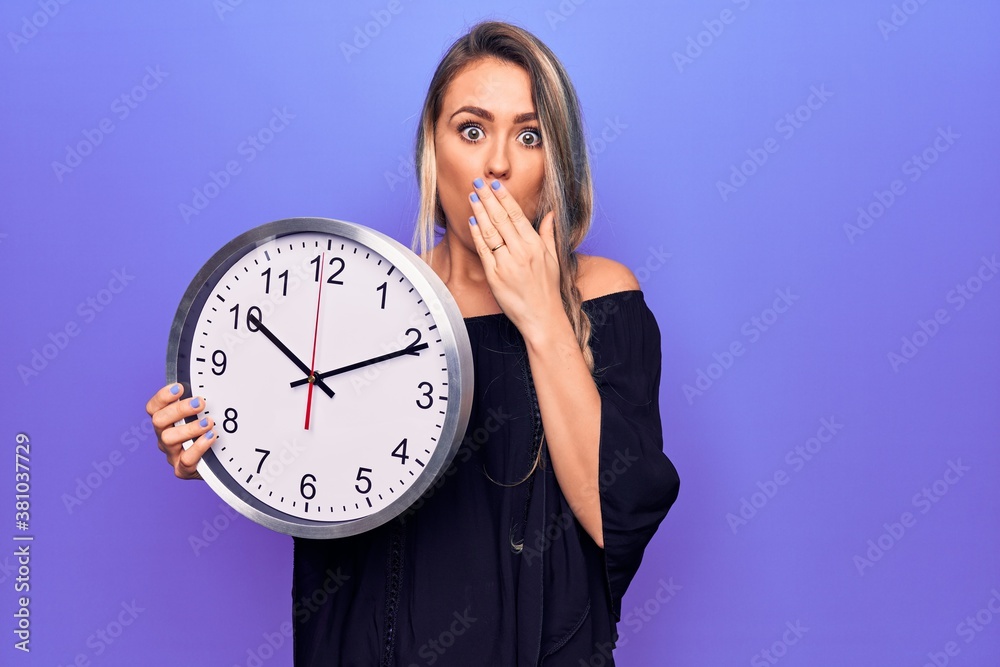 Young beautiful blonde woman doing countdown using big clock over purple background covering mouth with hand, shocked and afraid for mistake. Surprised expression Young beautiful blonde woman doing countdown using big clock over purple background covering mouth with hand, shocked and afraid for mistake. Surprised expression