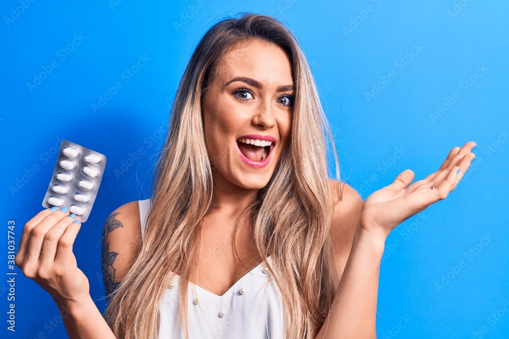 Young beautiful blonde woman holding medicine pills over isolated blue background celebrating achievement with happy smile and winner expression with raised hand Young beautiful blonde woman holding medicine pills over isolated blue background celebrating achievement with happy smile and winner expression with raised hand