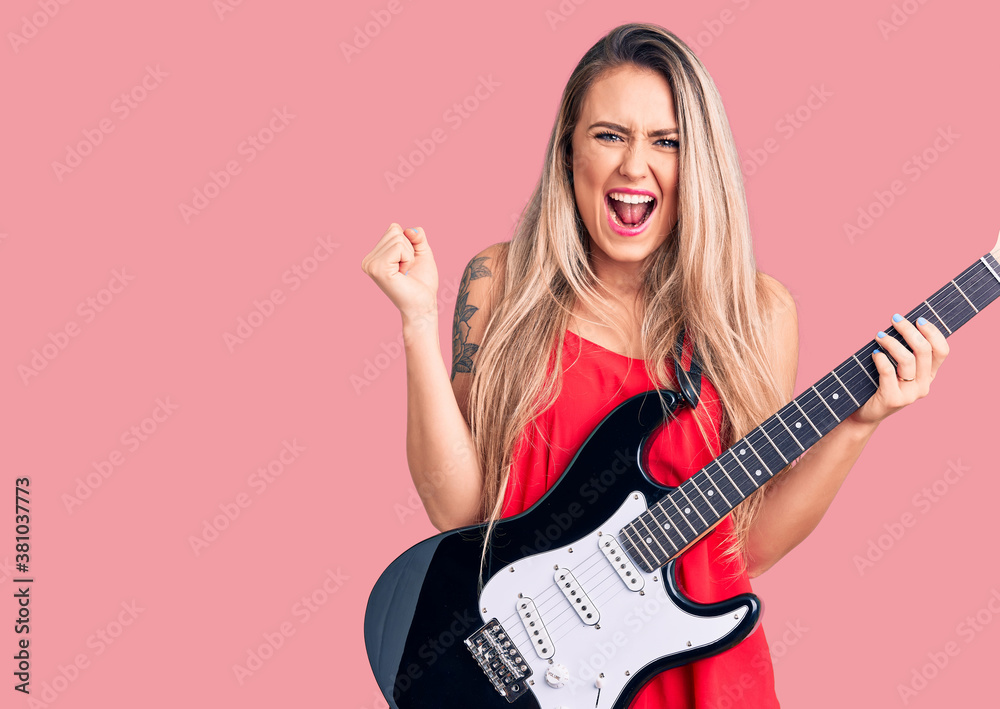Young beautiful blonde woman playing electric guitar screaming proud, celebrating victory and success very excited with raised arms Young beautiful blonde woman playing electric guitar screaming proud, celebrating victory and success very excited with raised arms