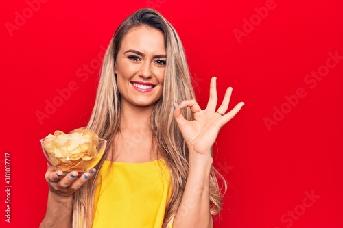 Young beautiful blonde woman holding bowl with potato chips over isolated red background doing ok sign with fingers  smiling friendly gesturing excellent symbol