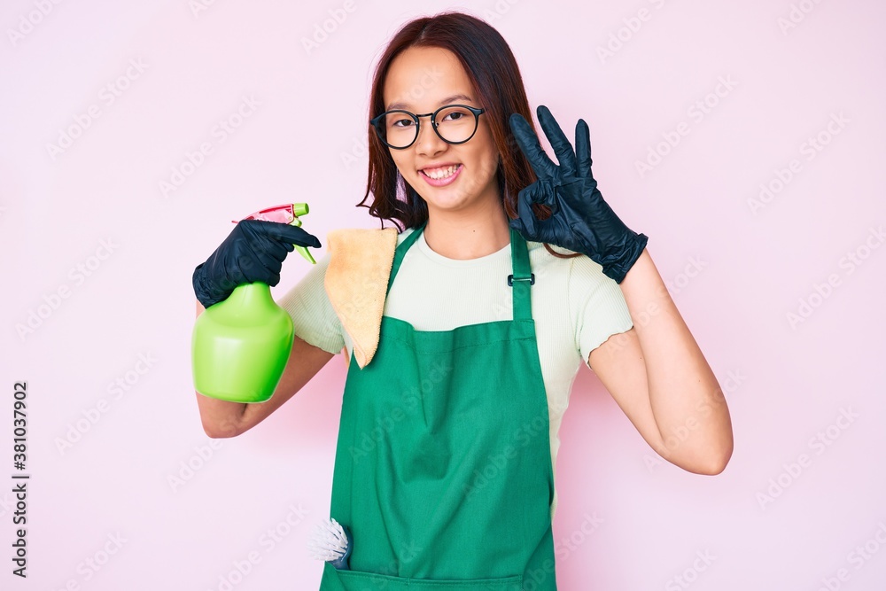 Young beautiful chinese girl wearing apron holding sprayer doing ok sign with fingers, smiling friendly gesturing excellent symbol Young beautiful chinese girl wearing apron holding sprayer doing ok sign with fingers, smiling friendly gesturing excellent symbol
