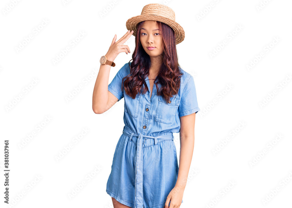 Young beautiful chinese girl wearing summer hat shooting and killing oneself pointing hand and fingers to head like gun, suicide gesture. Young beautiful chinese girl wearing summer hat shooting and killing oneself pointing hand and fingers to head like gun, suicide gesture.