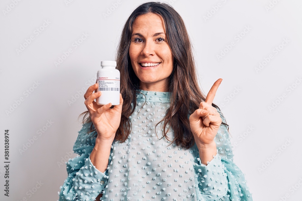 Young beautiful brunette woman holding jar of medicine pills over isolated white background smiling happy pointing with hand and finger to the side Young beautiful brunette woman holding jar of medicine pills over isolated white background smiling happy pointing with hand and finger to the side