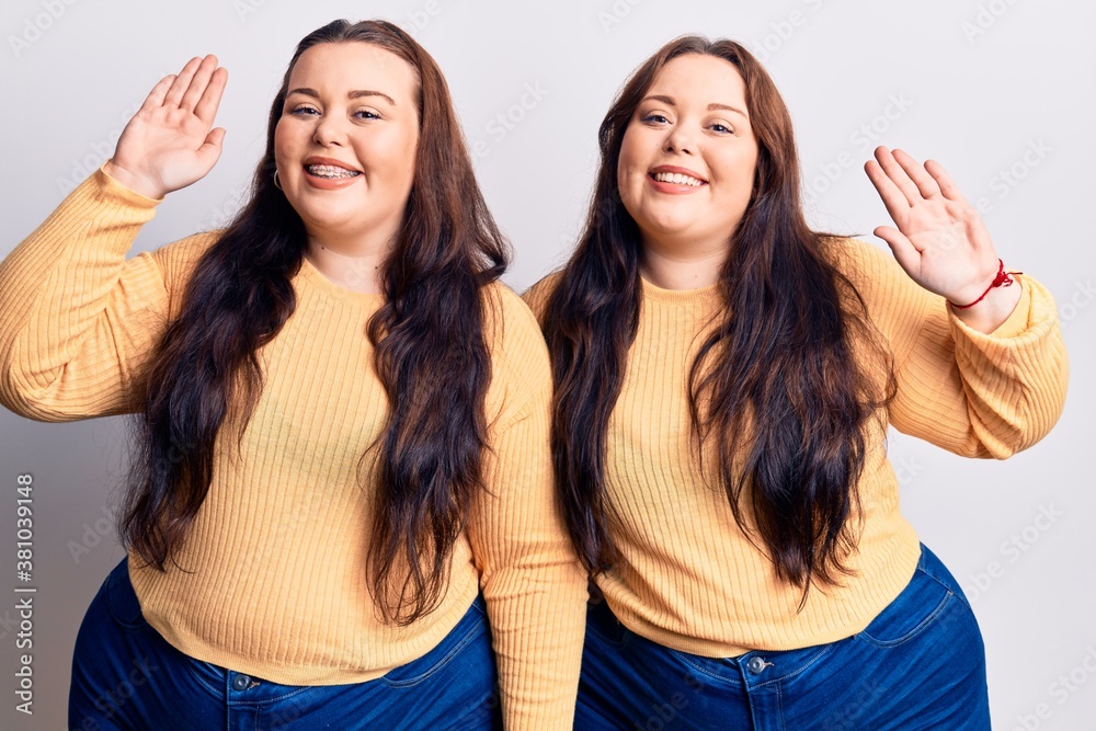 Young plus size twins wearing casual clothes waiving saying hello happy and smiling, friendly welcome gesture Young plus size twins wearing casual clothes waiving saying hello happy and smiling, friendly welcome gesture