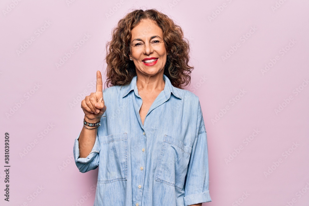Middle age beautiful woman wearing casual denim shirt standing over pink background showing and pointing up with finger number one while smiling confident and happy. Middle age beautiful woman wearing casual denim shirt standing over pink background showing and pointing up with finger number one while smiling confident and happy.
