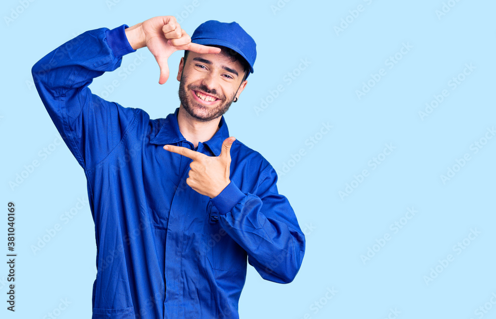 Young hispanic man wearing painter uniform smiling making frame with hands and fingers with happy face. creativity and photography concept.