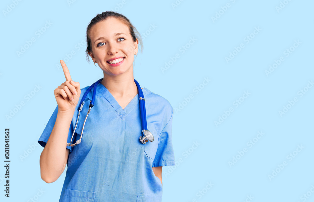 Young beautiful blonde woman wearing doctor uniform and stethoscope showing and pointing up with finger number one while smiling confident and happy. Young beautiful blonde woman wearing doctor uniform and stethoscope showing and pointing up with finger number one while smiling confident and happy.