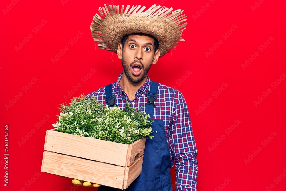 Young latin man wearing farmer hat and apron holding plant box pot scared and amazed with open mouth for surprise, disbelief face