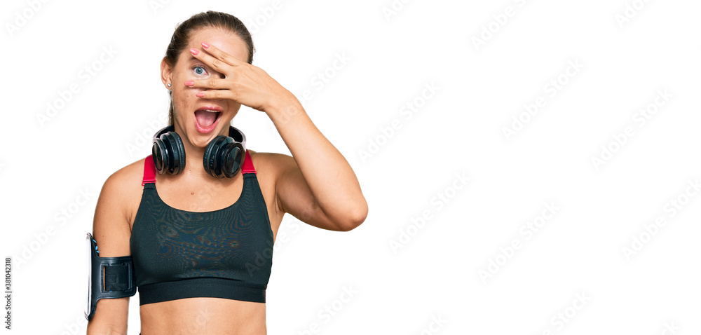 Beautiful blonde woman wearing gym clothes and using headphones peeking in shock covering face and eyes with hand, looking through fingers with embarrassed expression. Beautiful blonde woman wearing gym clothes and using headphones peeking in shock covering face and eyes with hand, looking through fingers with embarrassed expression.