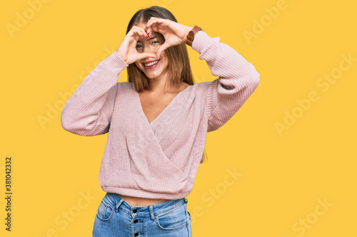 Beautiful blonde woman wearing casual winter pink sweater doing heart shape with hand and fingers smiling looking through sign