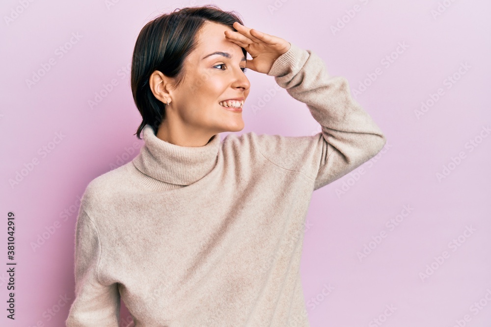 Young brunette woman with short hair wearing casual winter sweater very happy and smiling looking far away with hand over head. searching concept. Young brunette woman with short hair wearing casual winter sweater very happy and smiling looking far away with hand over head. searching concept.