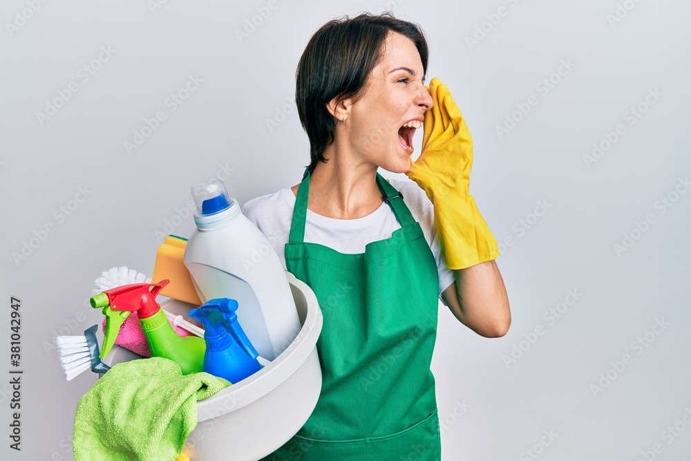 Young brunette woman with short hair wearing apron holding cleaning products shouting and screaming loud to side with hand on mouth. communication concept. Young brunette woman with short hair wearing apron holding cleaning products shouting and screaming loud to side with hand on mouth. communication concept.