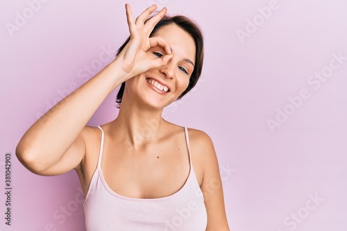 Young brunette woman with short hair over pink background smiling happy doing ok sign with hand on eye looking through fingers