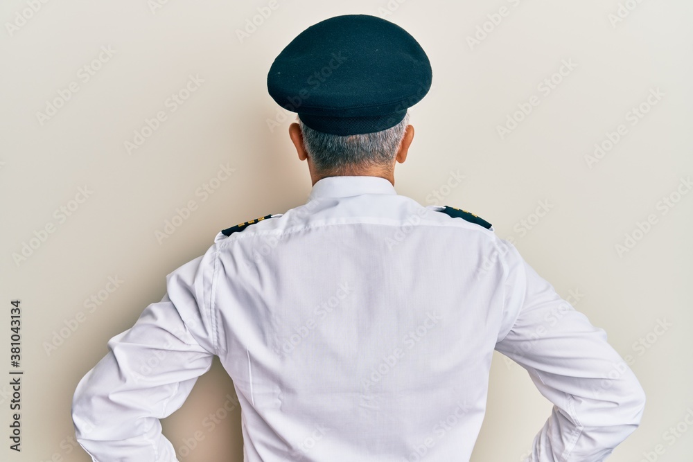 Handsome middle age mature man wearing airplane pilot uniform standing backwards looking away with arms on body Handsome middle age mature man wearing airplane pilot uniform standing backwards looking away with arms on body
