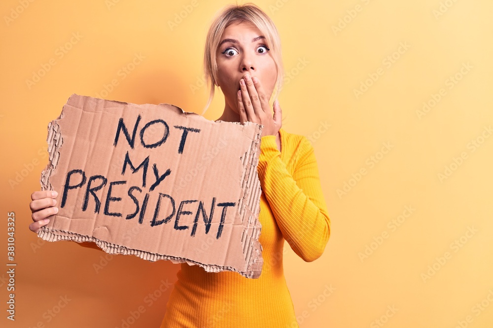 Young blonde woman asking for democracy holding banner with not my president message covering mouth with hand, shocked and afraid for mistake. Surprised expression Young blonde woman asking for democracy holding banner with not my president message covering mouth with hand, shocked and afraid for mistake. Surprised expression