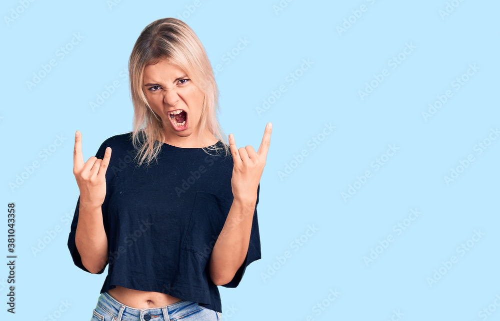 Young beautiful blonde woman wearing casual t-shirt shouting with crazy expression doing rock symbol with hands up. music star. heavy concept. Young beautiful blonde woman wearing casual t-shirt shouting with crazy expression doing rock symbol with hands up. music star. heavy concept.
