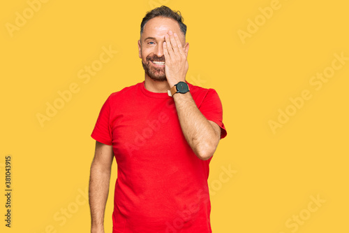 Handsome middle age man wearing casual red tshirt covering one eye with hand, confident smile on face and surprise emotion.