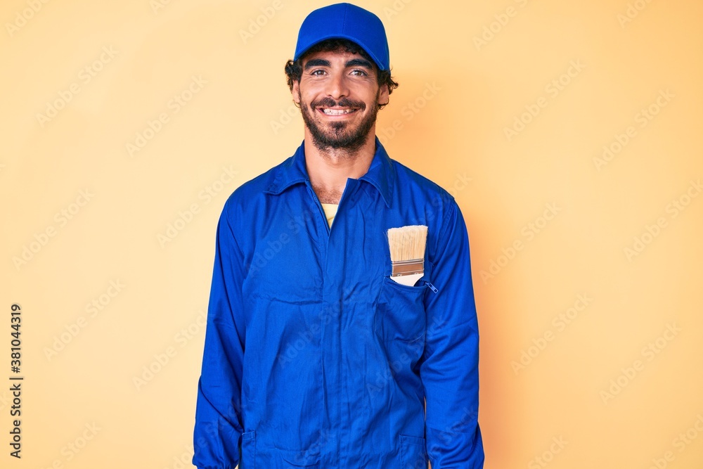 Handsome young man with curly hair and bear wearing builder jumpsuit uniform with a happy and cool smile on face. lucky person.
