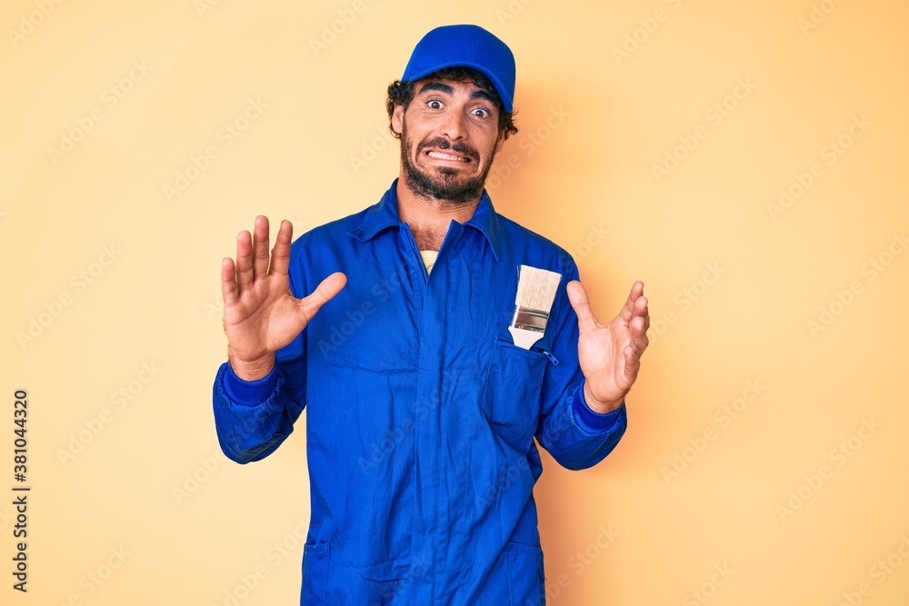 Handsome young man with curly hair and bear wearing builder jumpsuit uniform afraid and terrified with fear expression stop gesture with hands, shouting in shock. panic concept.