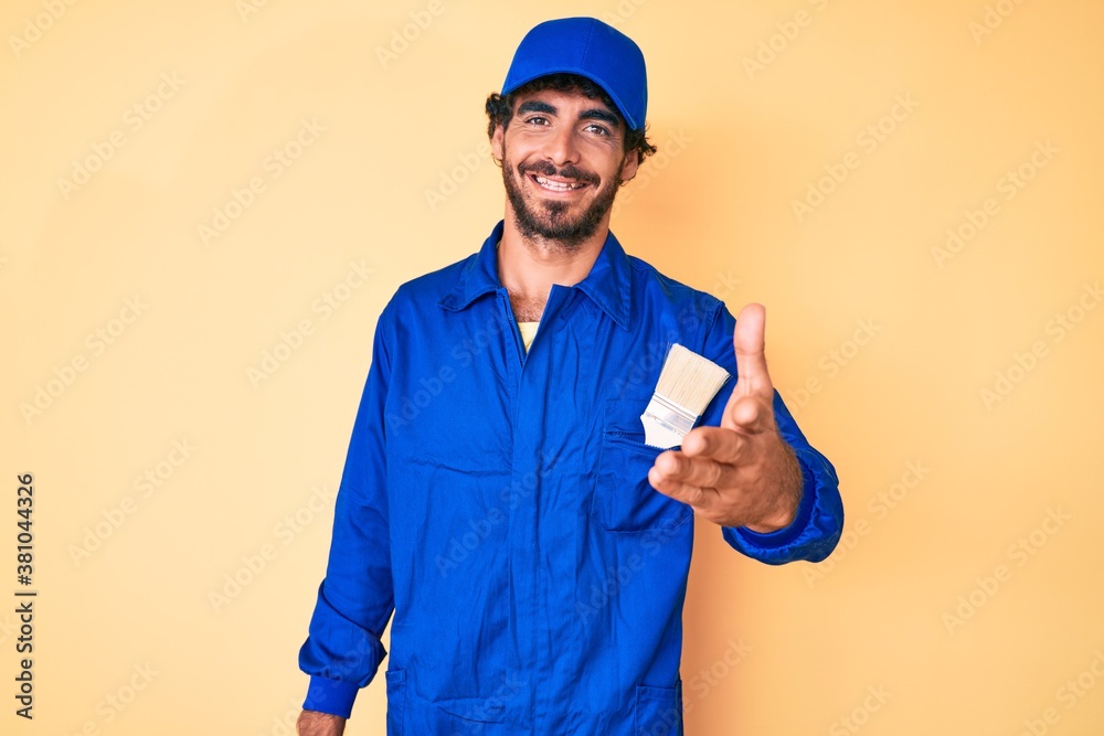 Handsome young man with curly hair and bear wearing builder jumpsuit uniform smiling friendly offering handshake as greeting and welcoming. successful business.