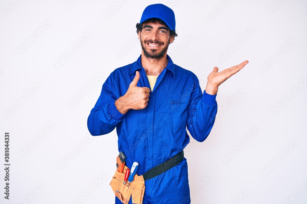 Handsome young man with curly hair and bear weaing handyman uniform showing palm hand and doing ok gesture with thumbs up, smiling happy and cheerful
