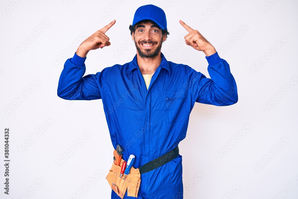 Handsome young man with curly hair and bear weaing handyman uniform smiling pointing to head with both hands finger, great idea or thought, good memory
