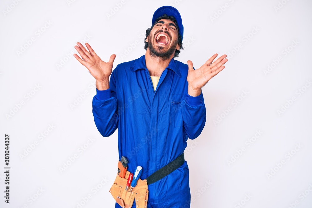 Handsome young man with curly hair and bear weaing handyman uniform celebrating mad and crazy for success with arms raised and closed eyes screaming excited. winner concept Handsome young man with curly hair and bear weaing handyman uniform celebrating mad and crazy for success with arms raised and closed eyes screaming excited. winner concept