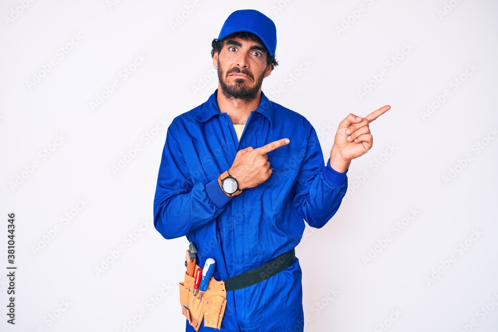 Handsome young man with curly hair and bear weaing handyman uniform pointing aside worried and nervous with both hands, concerned and surprised expression Handsome young man with curly hair and bear weaing handyman uniform pointing aside worried and nervous with both hands, concerned and surprised expression