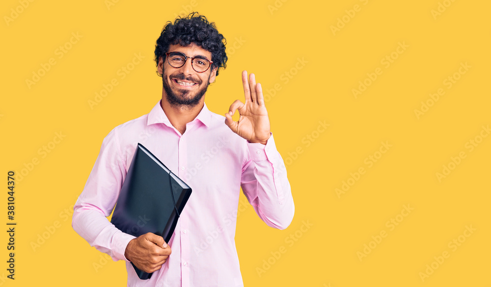 Handsome young man with curly hair and bear holding business folder doing ok sign with fingers, smiling friendly gesturing excellent symbol Handsome young man with curly hair and bear holding business folder doing ok sign with fingers, smiling friendly gesturing excellent symbol