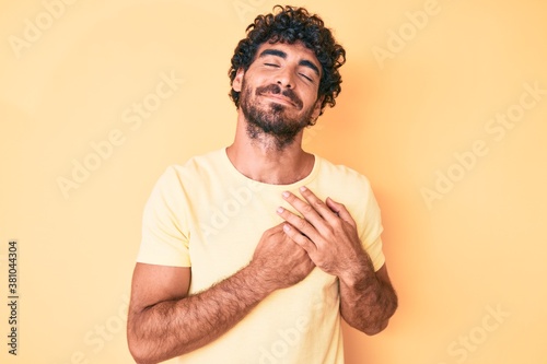 Foto Handsome young man with curly hair and bear wearing casual tshirt over yellow background smiling with hands on chest, eyes closed with grateful gesture on face