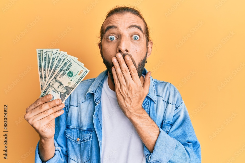 Attractive man with long hair and beard holding 20 dollars banknote covering mouth with hand, shocked and afraid for mistake. surprised expression Attractive man with long hair and beard holding 20 dollars banknote covering mouth with hand, shocked and afraid for mistake. surprised expression