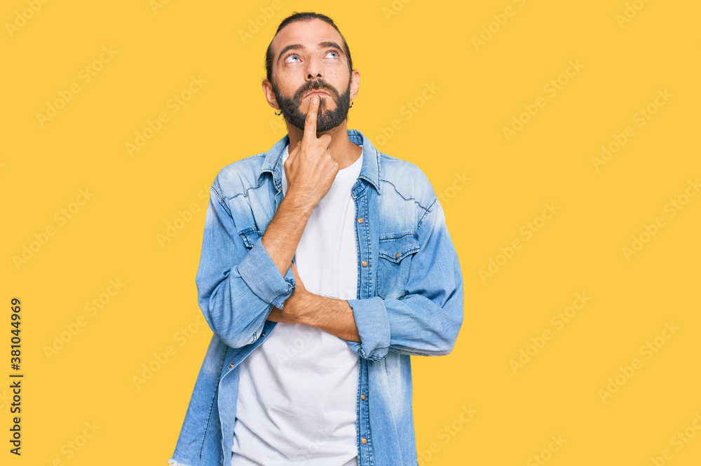Attractive man with long hair and beard wearing casual denim jacket thinking concentrated about doubt with finger on chin and looking up wondering Attractive man with long hair and beard wearing casual denim jacket thinking concentrated about doubt with finger on chin and looking up wondering