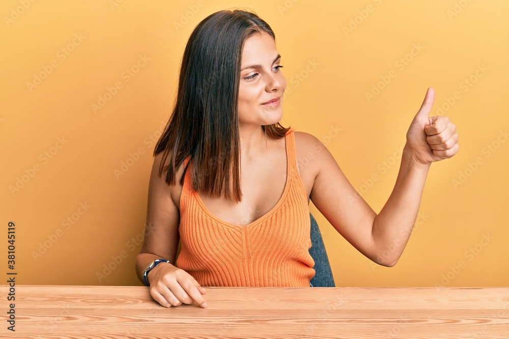 Young caucasian woman wearing casual clothes sitting on the table looking proud, smiling doing thumbs up gesture to the side Young caucasian woman wearing casual clothes sitting on the table looking proud, smiling doing thumbs up gesture to the side