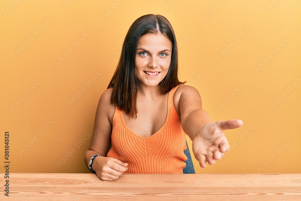 Young caucasian woman wearing casual clothes sitting on the table smiling cheerful offering palm hand giving assistance and acceptance. Young caucasian woman wearing casual clothes sitting on the table smiling cheerful offering palm hand giving assistance and acceptance.