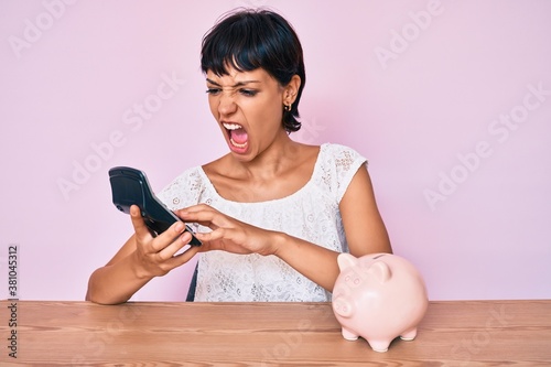 Beautiful brunettte woman caculating money savings angry and mad screaming frustrated and furious, shouting with anger. rage and aggressive concept. photo