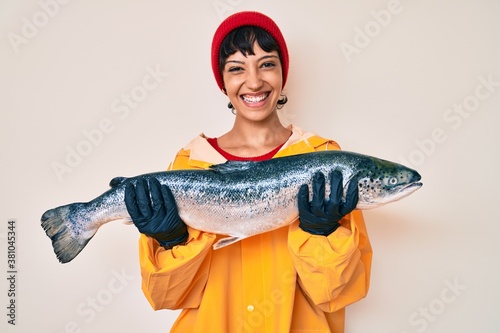 Beautiful brunettte fisher woman wearing raincoat holding fresh salmon smiling with a happy and cool smile on face. showing teeth. photo